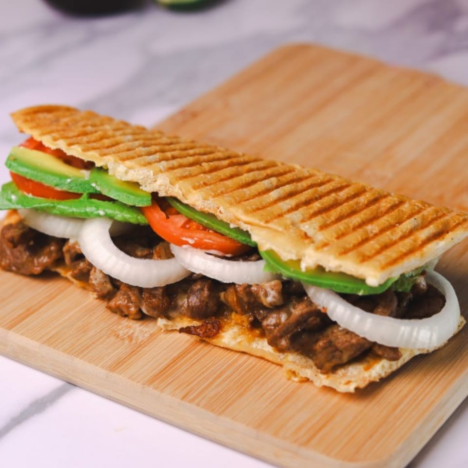 Amanecer Cocina y Cafe's breakfast panini with steak, onions, tomatoes, and avocado on a wooden board.