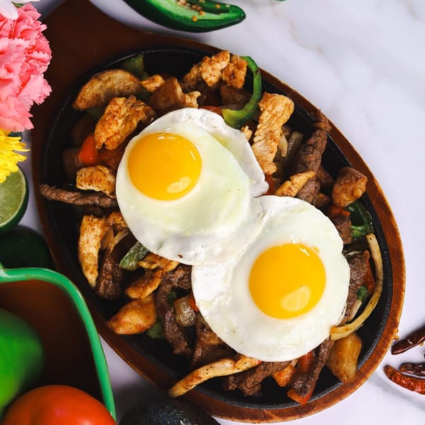 Sizzling fajitas with chicken, beef, peppers, onions, and two sunny-side-up eggs on a white surface.