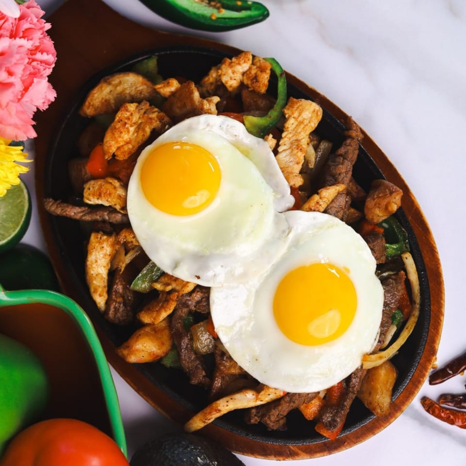 Sizzling fajitas with chicken, steak, peppers, onions, and two sunny-side-up eggs on a wooden platter.