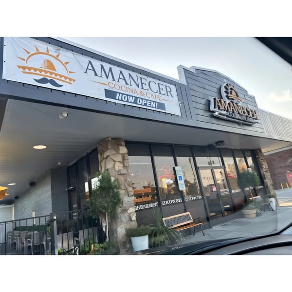Exterior of Amanecer Cocina y Cafe, a modern gray building with a "NOW OPEN!" banner and outdoor seating.