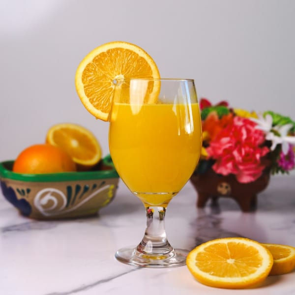 A glass of orange juice garnished with an orange slice, with oranges and flowers in the background.