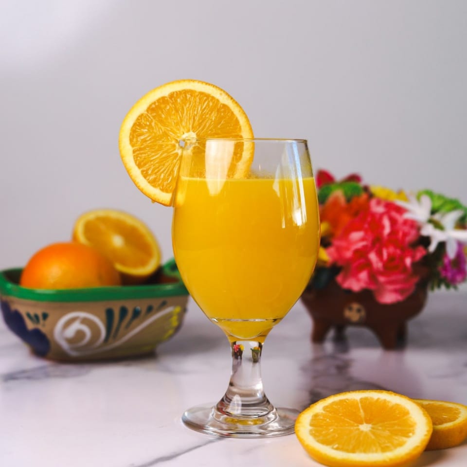 Fresh orange juice in a stemmed glass with an orange slice, whole oranges, and flowers.
