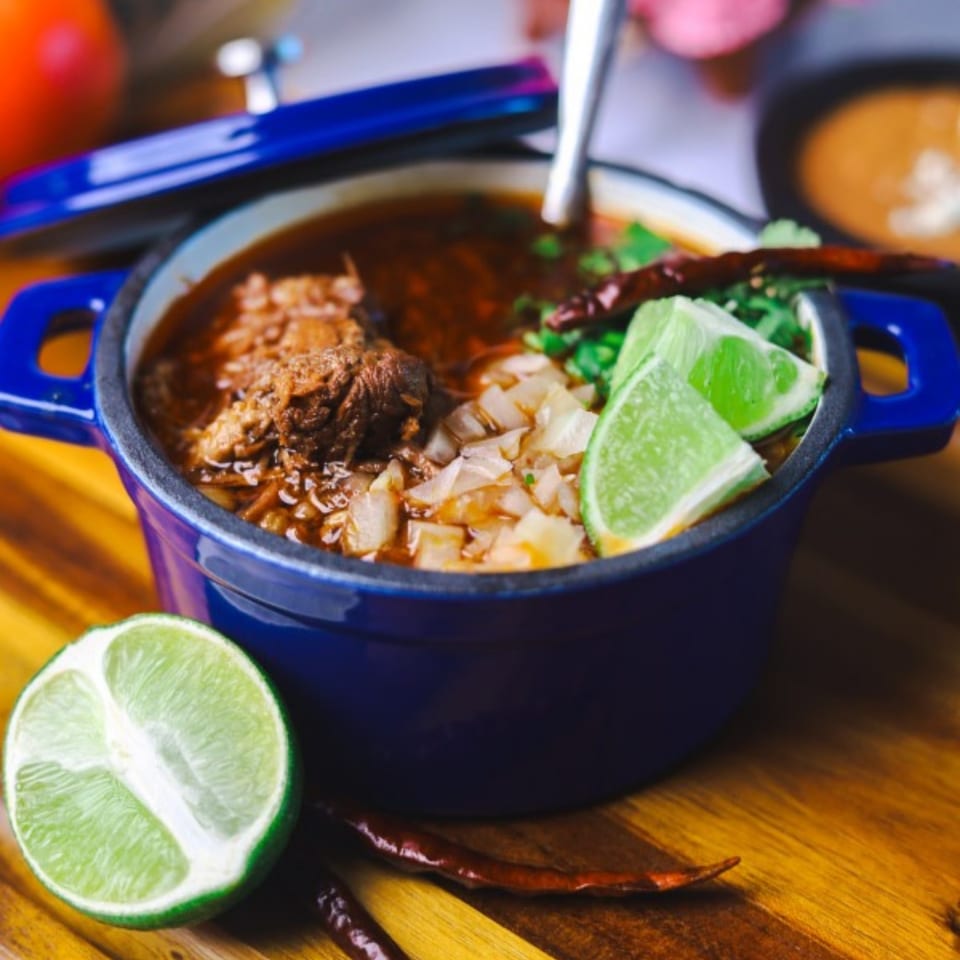 Amanecer Cocina y Cafe's birria in a blue pot, garnished with onions, cilantro, and lime.