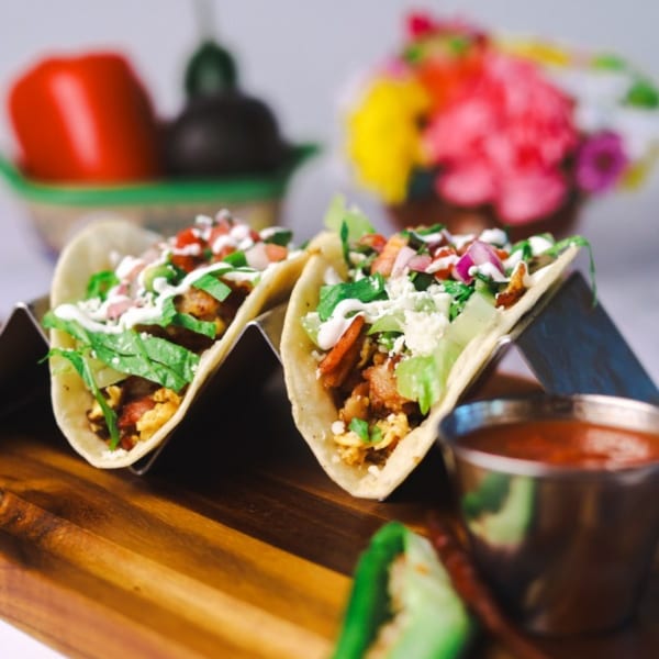 Two breakfast tacos with scrambled eggs, bacon, lettuce, pico de gallo, and crema, with salsa.