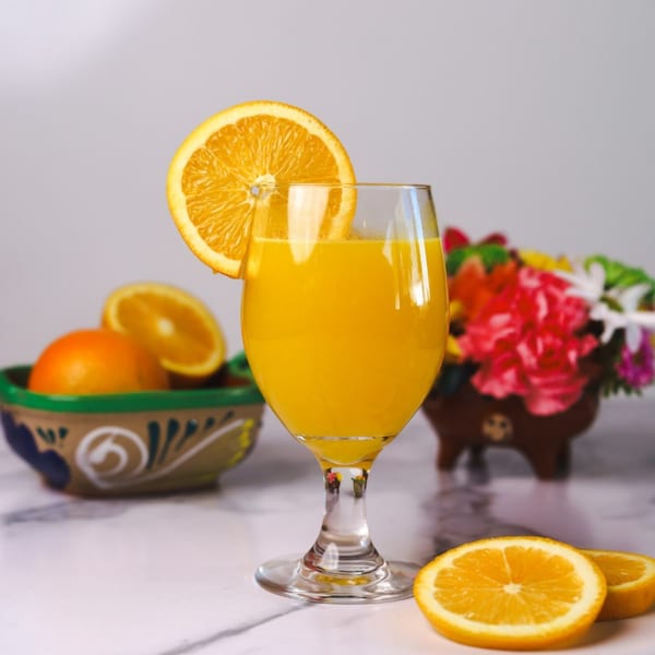 Fresh orange juice in a stemmed glass with an orange slice, whole oranges, and flowers.