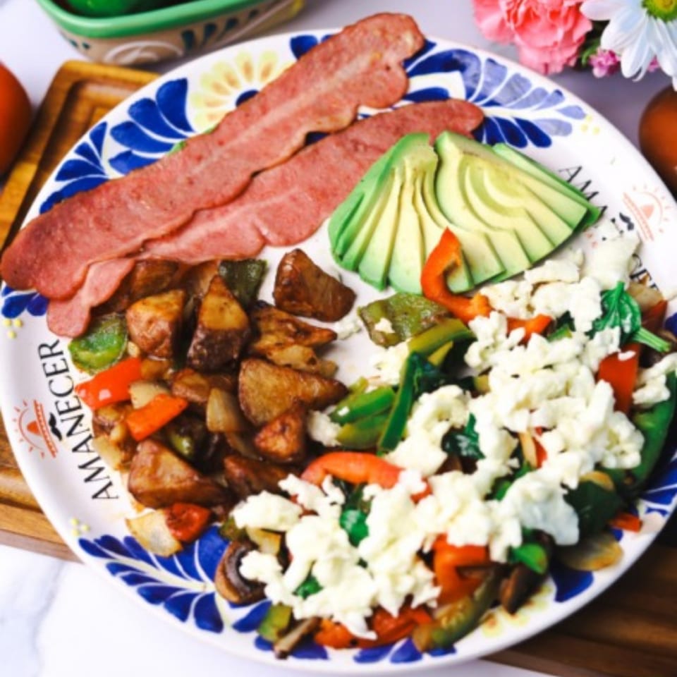Breakfast plate with bacon, avocado slices, roasted potatoes, and scrambled eggs with vegetables.